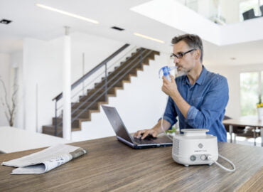 El hombre inhala con un sistema de inhalación mientras trabaja en una computadora portátil en la mesa