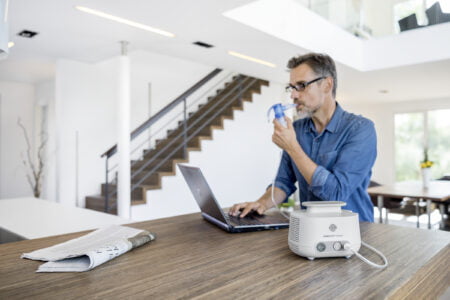 El hombre inhala con un sistema de inhalación mientras trabaja en una computadora portátil en la mesa
