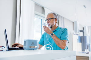 Un hombre mayor inhala con un sistema de inhalación mientras trabaja en una computadora portátil en la mesa